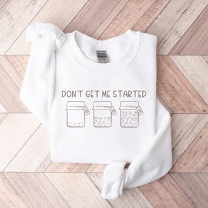 Peut inclure: Sweat-shirt blanc avec le texte "Don't get me started" et un graphique de trois bocaux avec des couvercles.