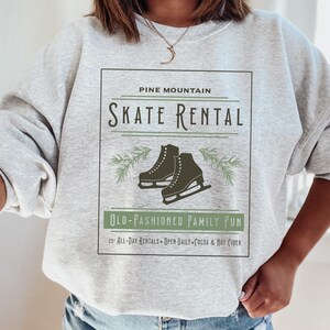 K&ouml;nnte beinhalten: Grauer Pullover mit einem grafischen Design eines Vintage-Schilds mit der Aufschrift "Pine Mountain Skate Rental" mit einem Paar Schlittschuhen und Kiefern&auml;sten. Der Text unter dem Schild lautet "Old-Fashioned Family Fun" und "25 All-Day Rentals - Open Daily - Cocoa & Hot Cider".