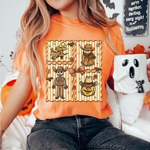 May include: Orange t-shirt with a Halloween design. The design features four square panels with cartoon illustrations of a witch, scarecrow, skeleton cowboy, and pumpkin cowboy. The shirt is made of a soft material.