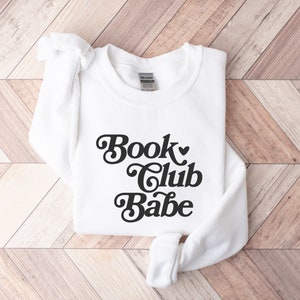 K&ouml;nnte beinhalten: Wei&szlig;er Pullover mit einem schwarzen Retro-Grafik, auf dem "Book Club Babe" mit einem Herzsymbol steht.