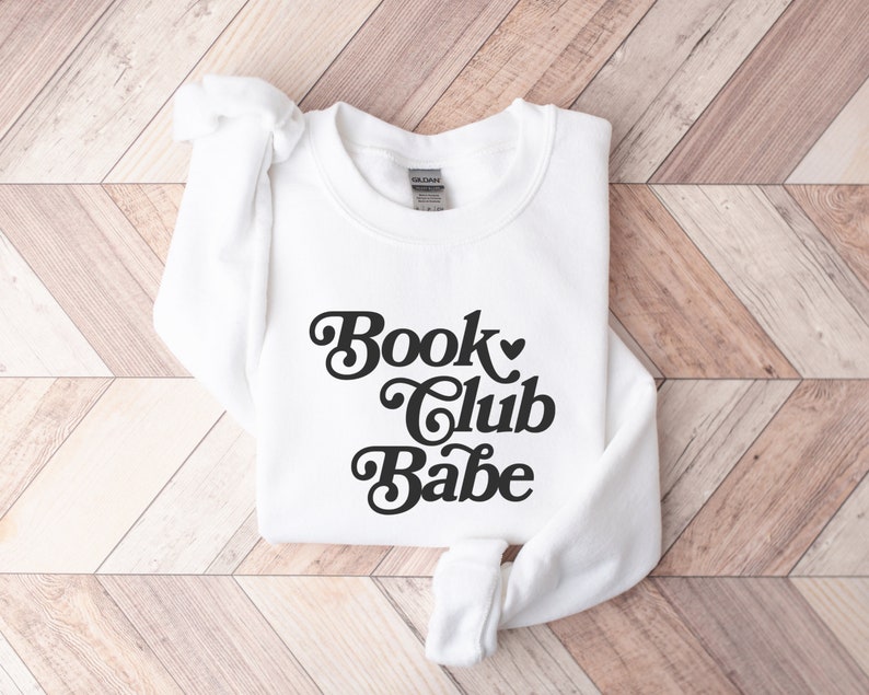 K&ouml;nnte beinhalten: Wei&szlig;er Pullover mit einem schwarzen Retro-Grafik, auf dem "Book Club Babe" mit einem Herzsymbol steht.