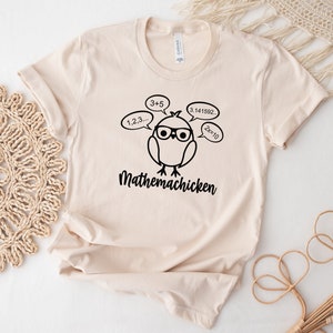 May include: A beige t-shirt with a black illustration of a chicken wearing glasses and thinking about math equations. The text "Mathemachicken" is printed below the chicken.
