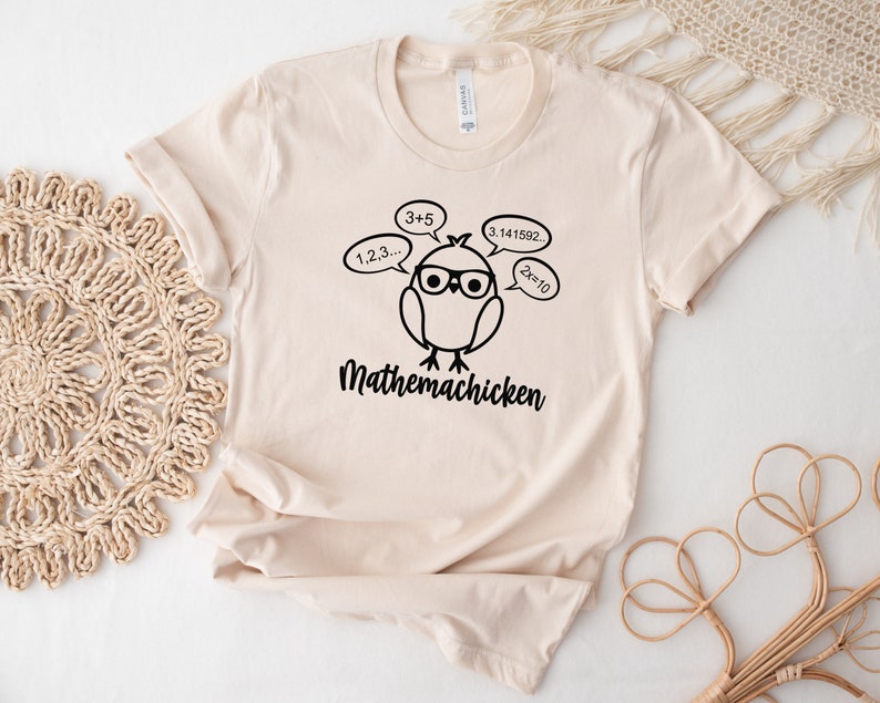 May include: A beige t-shirt with a black illustration of a chicken wearing glasses and thinking about math equations. The text "Mathemachicken" is printed below the chicken.