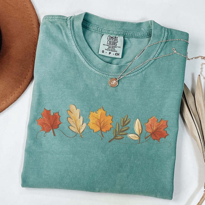 May include: A teal blue t-shirt with a graphic of five autumn leaves in shades of red, orange, yellow, and brown. The leaves are connected by a thin line. The shirt has a tag that reads "Comfort Colors" and "S - P - CH".