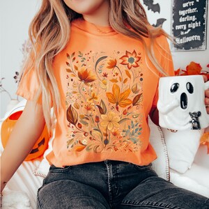 May include: Orange t-shirt featuring a floral design in shades of orange, yellow, and brown. The design includes various flowers and leaves. A white ghost-shaped mug is held in the hand. The background includes a Halloween sign.