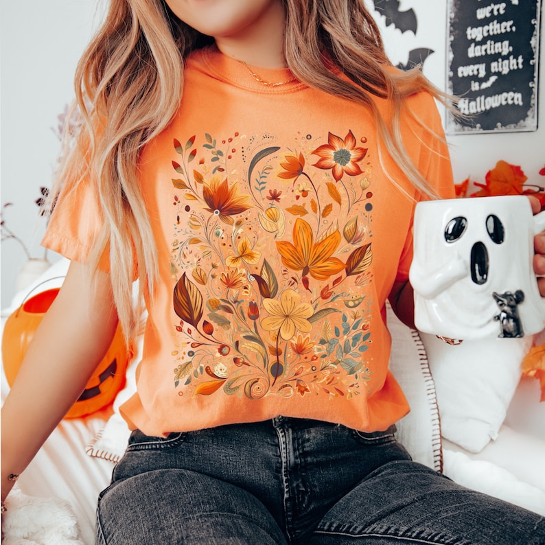May include: Orange t-shirt featuring a floral design in shades of orange, yellow, and brown. The design includes various flowers and leaves. A white ghost-shaped mug is held in the hand. The background includes a Halloween sign.