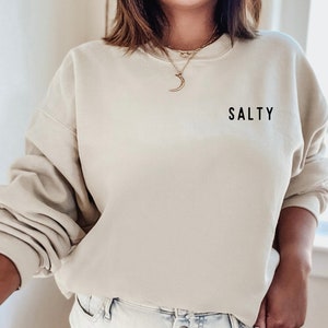 May include: A light beige sweatshirt with the word "SALTY" printed on the front in black. The sweatshirt has a crew neck and long sleeves.