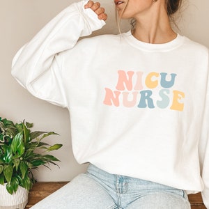 May include: White sweatshirt with a retro-style graphic that reads "NICU NURSE" in pastel pink, blue, and yellow.
