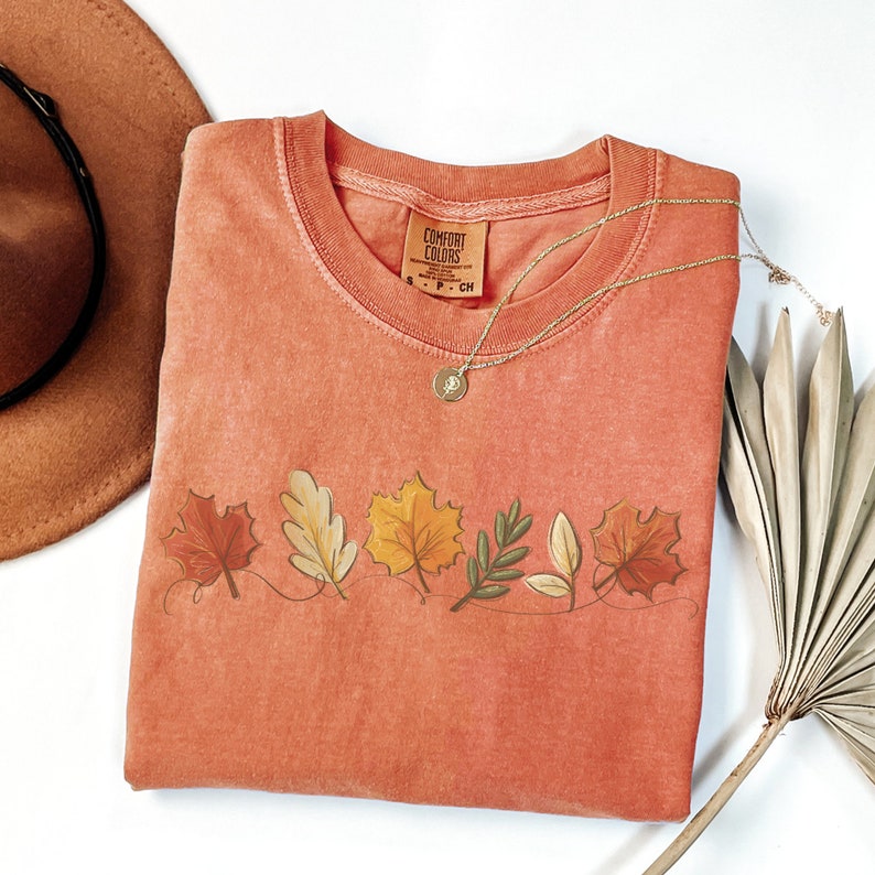 May include: A coral colored t-shirt with a row of fall leaves printed on the front. The leaves are in shades of orange, yellow, brown, and green. The shirt has a crew neck and short sleeves. The shirt is made by Comfort Colors.