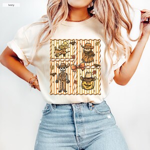 May include: Ivory t-shirt with a four-panel Halloween design. Each panel displays a different character: a witch on a broom, a scarecrow, a skeleton cowboy, and a pumpkin wearing a cowboy hat. The design includes bats and leaves, with a striped background.