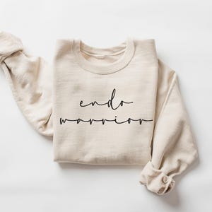 May include: A cream-colored sweatshirt with the words "endo warrior" in black script. The long sleeves are cuffed, and the crew neck is visible. The sweatshirt is made of a soft material and is laid flat on a white surface.