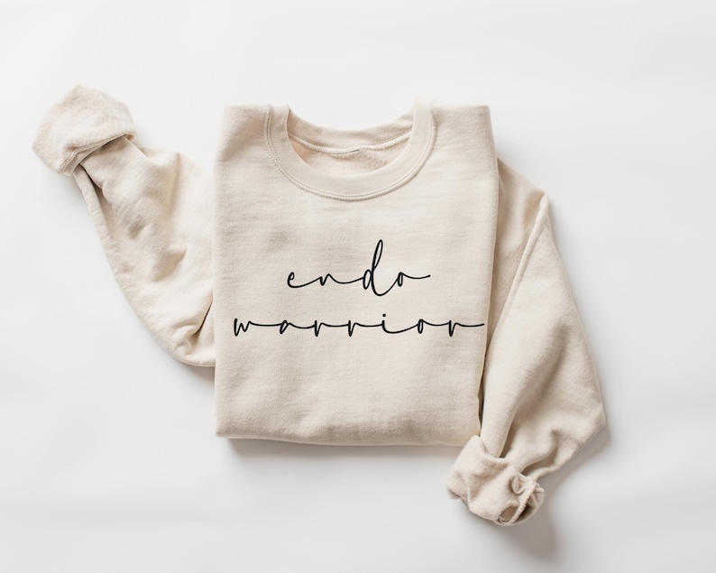 May include: A cream-colored sweatshirt with the words "endo warrior" in black script. The long sleeves are cuffed, and the crew neck is visible. The sweatshirt is made of a soft material and is laid flat on a white surface.