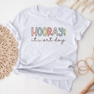 May include: A grey t-shirt with the text "HOORAY! it's art day" in colourful letters.
