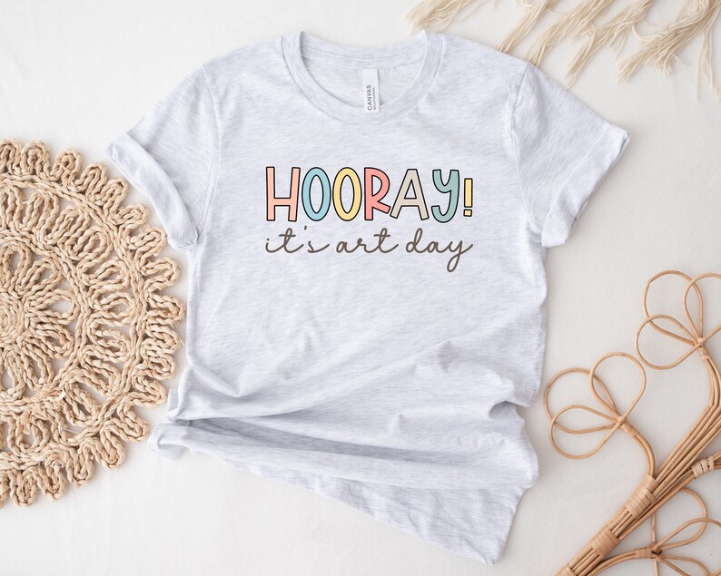 May include: A grey t-shirt with the text "HOORAY! it's art day" in colourful letters.