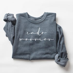May include: A dark gray sweatshirt with the words "endo warrior" in white script. The long-sleeved shirt is folded, with the sleeves cuffed. The background is plain white.