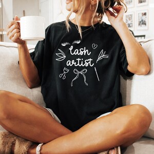 May include: A woman wearing a black t-shirt with the text "lash artist" in white lettering. The shirt also features a stylized eyelash, a heart, a flower, scissors, a glass of wine, a ribbon, and a needle.