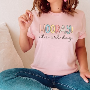 May include: A pink t-shirt with the text "HOORAY! it's art day" in colourful letters.