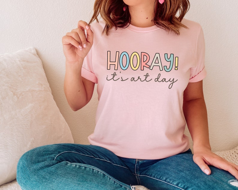 May include: A pink t-shirt with the text "HOORAY! it's art day" in colourful letters.