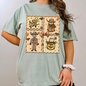 May include: Sage green t-shirt with a four-panel design featuring Halloween characters. Each panel has a vertical striped background with a witch, scarecrow, skeleton, and pumpkin wearing cowboy hats. Autumn leaves and bats are also included.
