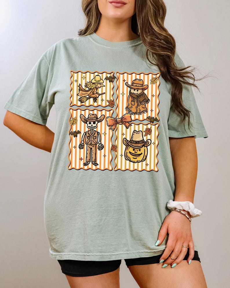 May include: Sage green t-shirt with a four-panel design featuring Halloween characters. Each panel has a vertical striped background with a witch, scarecrow, skeleton, and pumpkin wearing cowboy hats. Autumn leaves and bats are also included.