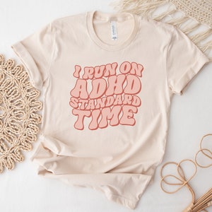 May include: A beige t-shirt with the text "I run on ADHD standard time" in pink retro-style lettering.