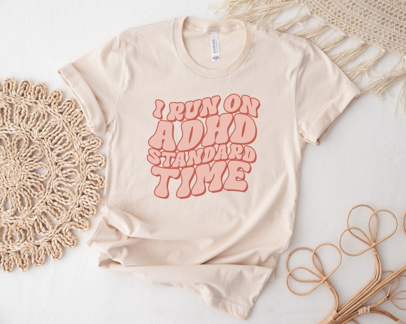 May include: A beige t-shirt with the text "I run on ADHD standard time" in pink retro-style lettering.