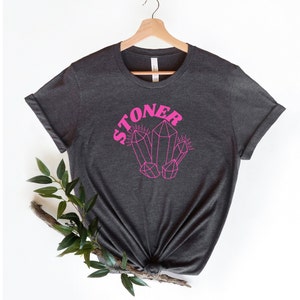May include: A dark gray t-shirt with a pink graphic of crystals and the word "STONER" in pink.