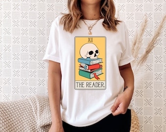 May include: White t-shirt with a graphic of a tarot card. The card is yellow with a skull on top of a stack of books. The card reads "XII THE READER."