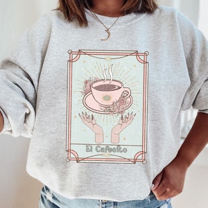 May include: Gray sweatshirt with a graphic of a coffee cup in a vintage tarot card style. The card reads "El Cafeito".