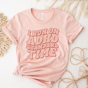 May include: A pink t-shirt with the text "I run on ADHD standard time" in a retro font style.