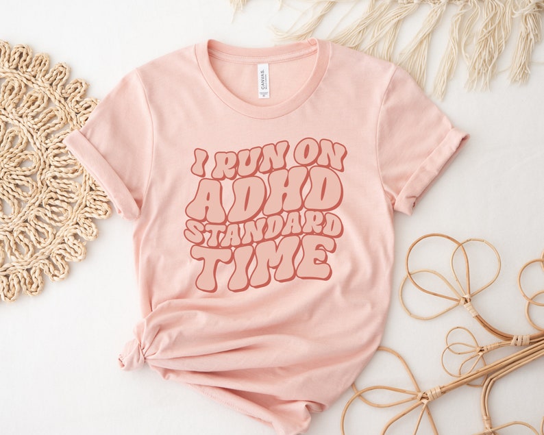 May include: A pink t-shirt with the text "I run on ADHD standard time" in a retro font style.