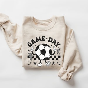 May include: A beige sweatshirt with a black and white graphic of a soccer ball with arms and legs. The graphic says "GAME DAY" in a retro font.