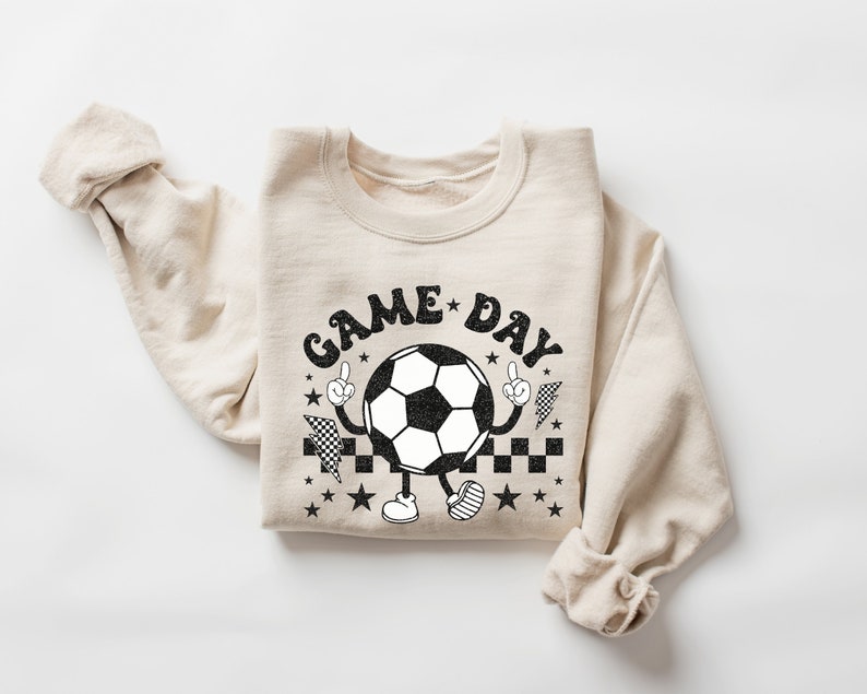 May include: A beige sweatshirt with a black and white graphic of a soccer ball with arms and legs. The graphic says "GAME DAY" in a retro font.