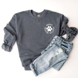 May include: A dark gray sweatshirt with a white paw print and the text "Adopt - Foster - Rescue" around it. The sweatshirt is paired with light blue jeans.