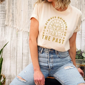 K&ouml;nnte beinhalten: Ein beiges T-Shirt mit einer goldenen Grafik eines &auml;gyptischen Pharaos und dem Text "I'm always bringing up the past".