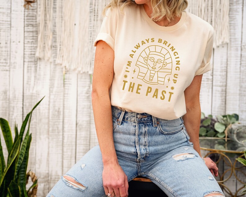 K&ouml;nnte beinhalten: Ein beiges T-Shirt mit einer goldenen Grafik eines &auml;gyptischen Pharaos und dem Text "I'm always bringing up the past".