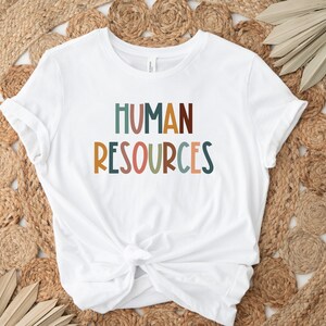 May include: White t-shirt with the text "HUMAN RESOURCES" in colorful, stylized letters.