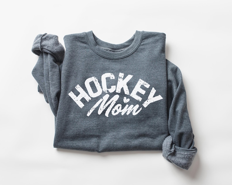 May include: A dark gray sweatshirt with the text "HOCKEY MOM" in white with a distressed look. The sweatshirt is folded and laid flat on a white surface.