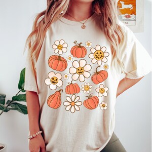 K&ouml;nnte beinhalten: Cremefarbenes T-Shirt mit einem Retro-Design aus orangefarbenen K&uuml;rbissen und wei&szlig;en G&auml;nsebl&uuml;mchen mit l&auml;chelnden Gesichtern. Das Shirt hat eine lockere Passform und kurze &Auml;rmel, ideal f&uuml;r den Herbst. Die Grafik hat einen Vintage-Look.