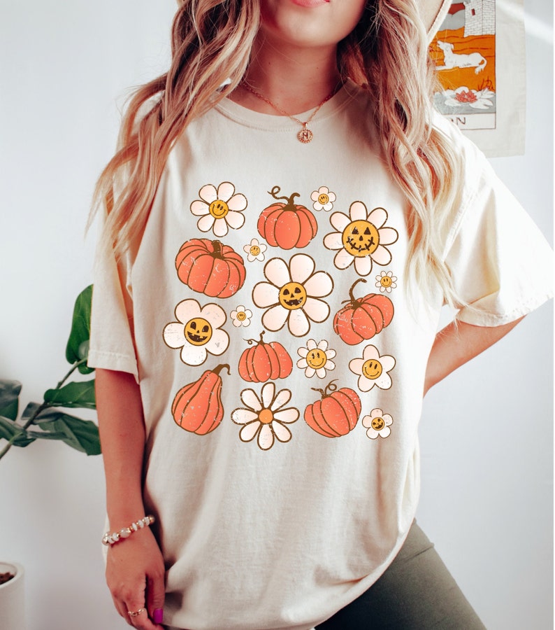 K&ouml;nnte beinhalten: Cremefarbenes T-Shirt mit einem Retro-Design aus orangefarbenen K&uuml;rbissen und wei&szlig;en G&auml;nsebl&uuml;mchen mit l&auml;chelnden Gesichtern. Das Shirt hat eine lockere Passform und kurze &Auml;rmel, ideal f&uuml;r den Herbst. Die Grafik hat einen Vintage-Look.