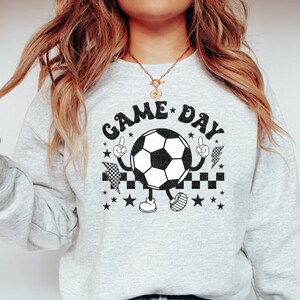 May include: A gray sweatshirt with the text "GAME DAY" in black letters. A cartoon soccer ball with arms and legs is wearing a checkered racing flag and pointing with one finger.