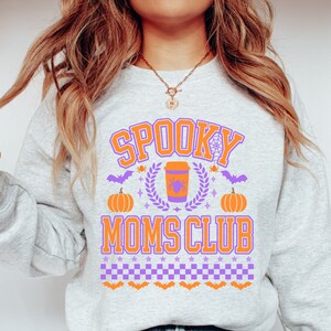 May include: A gray sweatshirt with the text "Spooky Moms Club" in purple and orange. The design includes a coffee cup with a spider, pumpkins, bats, and stars.