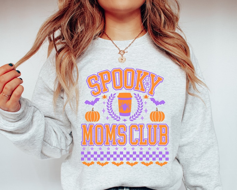 May include: A gray sweatshirt with the text "Spooky Moms Club" in purple and orange. The design includes a coffee cup with a spider, pumpkins, bats, and stars.