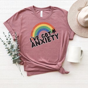 May include: A mauve t-shirt with a rainbow and a star graphic. The text "I've got anxiety" is printed in black on the shirt.