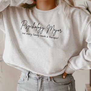 以下が含まれることがあります： 黒い文字で「Psychology Major cuz every family needs a therapist」と書かれた灰色のスウェットシャツです。