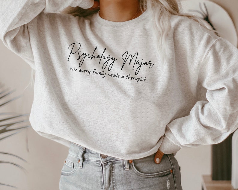 以下が含まれることがあります： 黒い文字で「Psychology Major cuz every family needs a therapist」と書かれた灰色のスウェットシャツです。