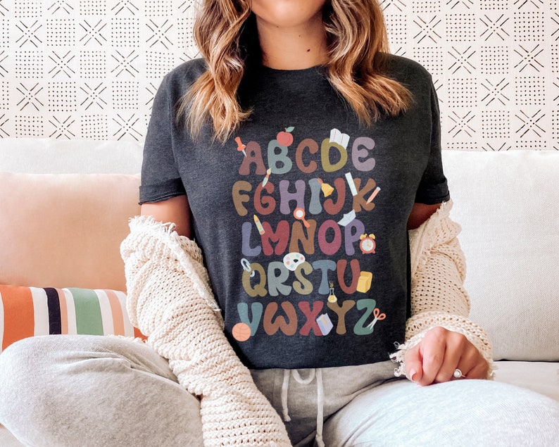 Pode incluir: Uma camiseta cinza escura com um design de alfabeto colorido que apresenta ilustra&ccedil;&otilde;es com tema escolar. As letras est&atilde;o em uma fonte arredondada e divertida, e cada letra est&aacute; emparelhada com uma pequena imagem como uma ma&ccedil;&atilde;, um l&aacute;pis, um pincel, um rel&oacute;gio e uma tesoura.