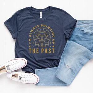 K&ouml;nnte beinhalten: Ein marineblaues T-Shirt mit einer goldenen Grafik eines &auml;gyptischen Pharaos im Kreis. Der Text "I'm always bringing up the past" ist um den Kreis herum geschrieben.