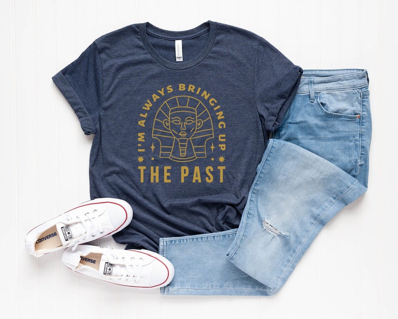 K&ouml;nnte beinhalten: Ein marineblaues T-Shirt mit einer goldenen Grafik eines &auml;gyptischen Pharaos im Kreis. Der Text "I'm always bringing up the past" ist um den Kreis herum geschrieben.