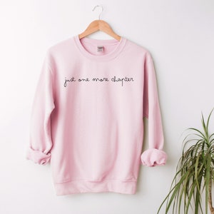 May include: A pink sweatshirt with the text "just one more chapter" in black script.